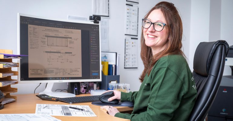 Person at a desk with a computer screen showing technical drawings, using a mouse.